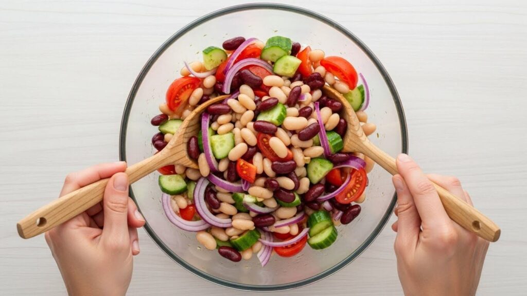 Hands tossing ingredients for 3-bean salad dressing in a glass bowl