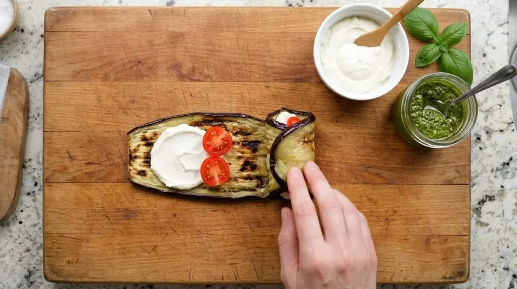 Step-by-step process of assembling eggplant rollatini by spreading ricotta filling and cherry tomatoes on a roasted eggplant slice on a wooden board.