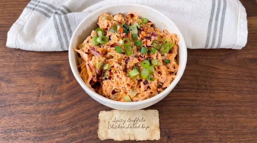 A white bowl filled with spicy buffalo chicken salad dip garnished with green onions and red pepper flakes on a wooden table.