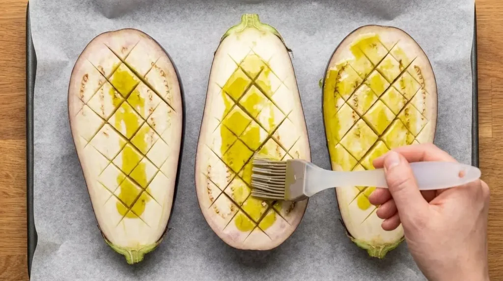 Brushing extra virgin olive oil into deep diamond scored cuts on raw eggplant halves before roasting.