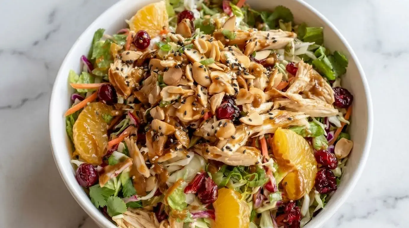 Large white bowl filled with Mediterranean Style Asian Chicken Salad topped with shredded chicken, cabbage, mandarin oranges, toasted almonds, peanuts, and black sesame seeds.