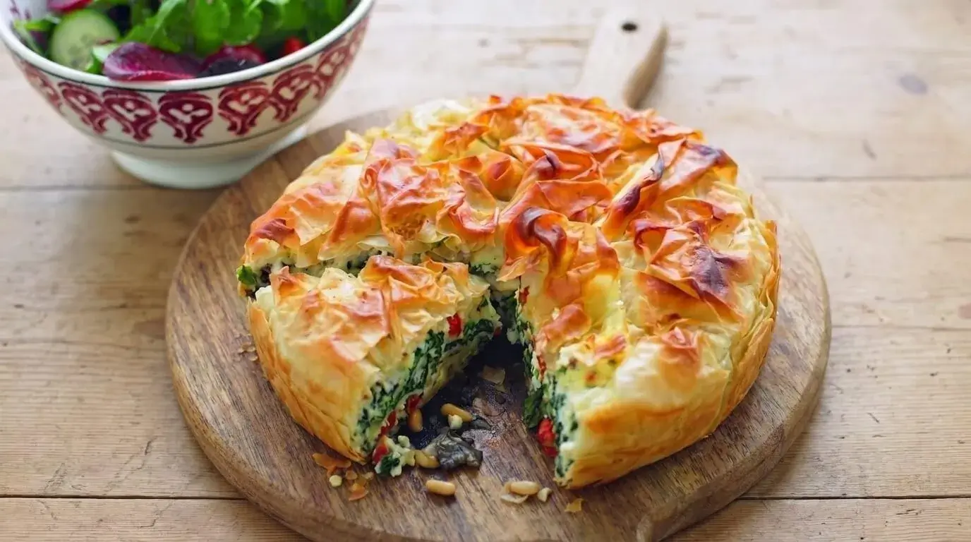 A sliced golden-brown one-pan feta filo tart on a wooden board showing spinach, feta, and sun-dried tomato filling.