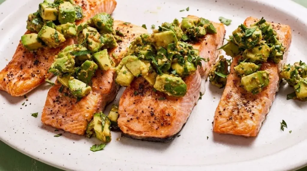 Broiled salmon fillets topped with fresh green avocado gremolata salad on a white serving plate.